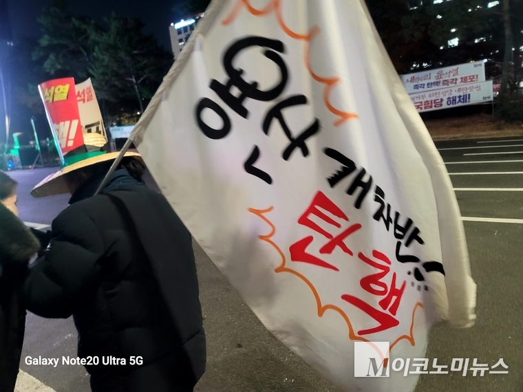 한 참가자가 "내란수괴 윤석열 탄핵"이라고 적힌 피켓을 둥그렇게 말아서 붙인 모자와 "용산 개차반! 탄핵"이라고 적인 깃발을 들고 있다.